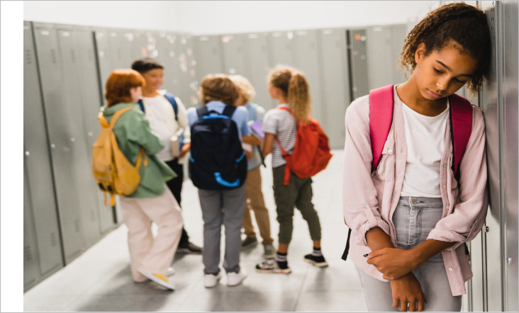 A young girl with a backpack leans against a locker, looking sad and isolated, while a group of children talk together in the background. The image highlights feelings of loneliness and exclusion in a school setting.