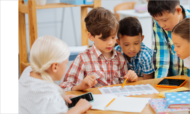 A group of young students working together at a classroom table, using pencils and calculators. The image highlights teamwork, collaboration, and engaged learning.