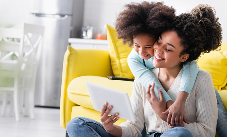 A mother and her young daughter share a moment at home. The mother, sitting on the floor with a tablet in her hands, smiles as her daughter hugs her from behind. They are in a cozy, living room with a bright yellow couch in the background.