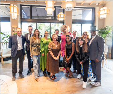 A diverse group of people poses together in a well-lit, modern indoor space. They are dressed in a mix of formal and stylish outfits, smiling at the camera. The background features large windows, hanging lights, and greenery.