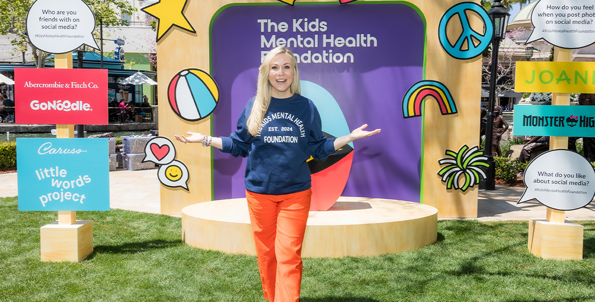 Actress and mental health advocate Ashley Eckstein stands smiling at a Kids Mental Health Foundation event, surrounded by colorful signage and brand partners supporting youth emotional wellness.
