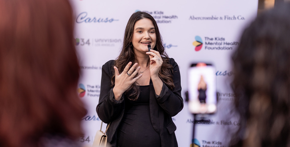 Sam DeRosa performs live with a microphone while a guitarist accompanies her, during an evening event supporting youth mental health hosted by The Kids Mental Health Foundation.