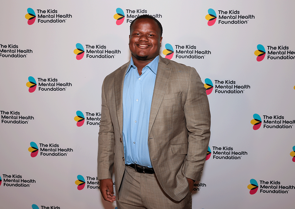 Tyler Davis poses at a The Kids Mental Health Foundation event in front of a backdrop featuring the organization’s logo.