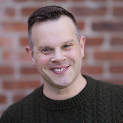 Headshot of Andrew Levitt, known professionally as Nina West, smiling in front of a brick wall.