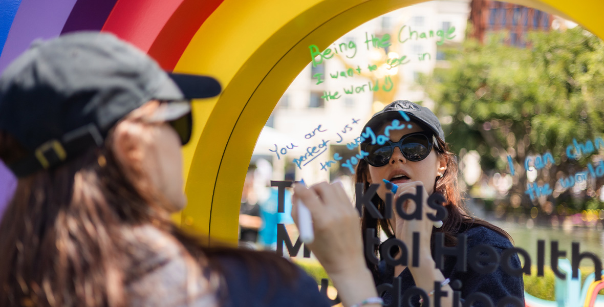 Alexandra Daddario writes encouraging messages on a reflective board at a Kids Mental Health Foundation event, participating in a community activity promoting youth confidence and positivity.
