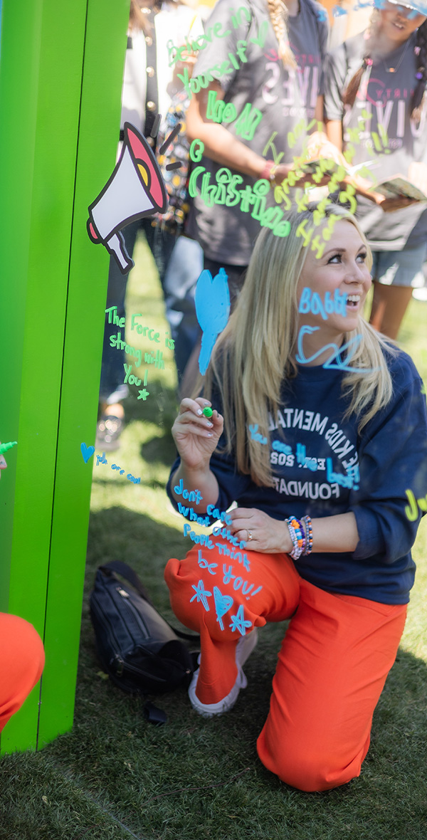 Ashley Eckstein kneels beside a mirror wall writing uplifting messages during a Kids Mental Health Foundation activity, encouraging kids to express themselves and build confidence through creativity.