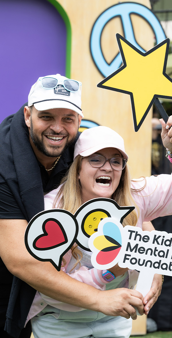 Daniel Franzese poses with a smiling supporter at a Kids Mental Health Foundation event, holding playful photo booth props with hearts, stars, and the foundation's logo to celebrate youth mental health.