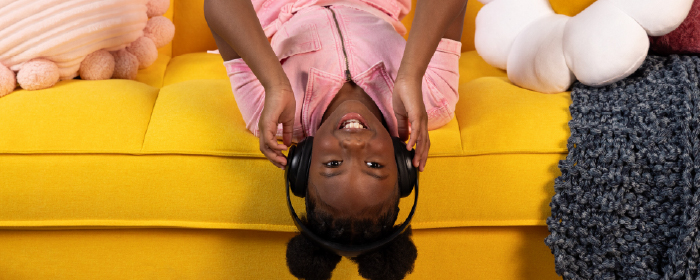 Smiling girl wearing headphones lounges upside down on a bright yellow couch, representing joy, creativity, and emotional wellness supported by The Kids Mental Health Foundation.