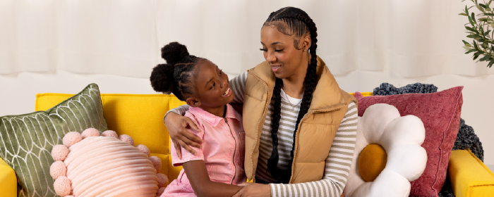 Mother and daughter sit closely on a yellow couch with playful pillows, showcasing strong family bonds and the importance of community partnerships in mental wellness."