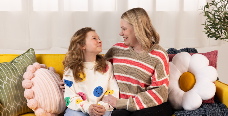 Mother and daughter sit on a yellow couch surrounded by colorful pillows, smiling at each other, symbolizing a strong parent-child bond and emotional support at home.