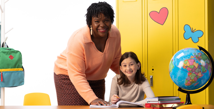 Teacher and student smile together at a desk with books and a globe, in front of bright yellow lockers decorated with a heart and butterfly, representing positive learning and classroom support.