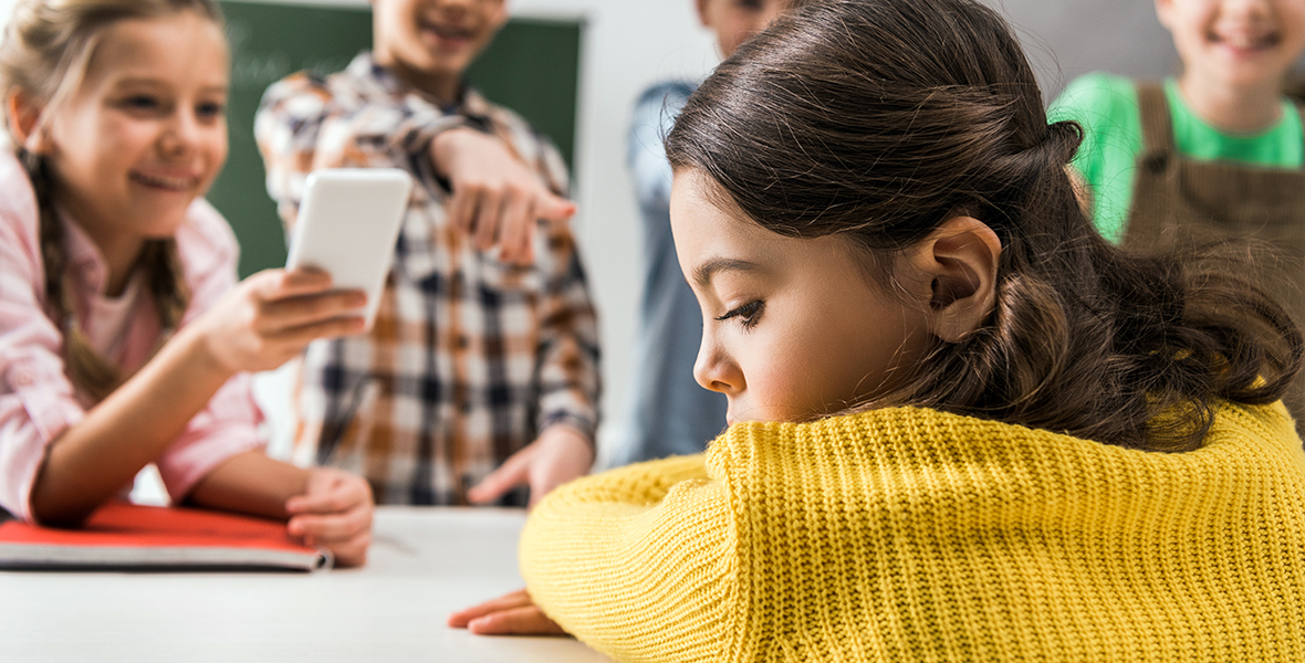 Sad young girl being bullied by classmates in school, with peers pointing and using a smartphone to take photos. Raising awareness about cyberbullying, social exclusion, and the impact on children's mental health.