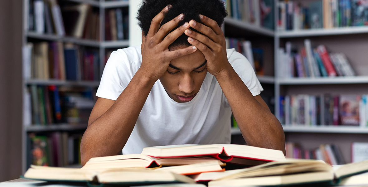 Stressed student holding his head in a library, overwhelmed with studying. Academic pressure and mental health challenges among youth, highlighting the importance of stress management and support.