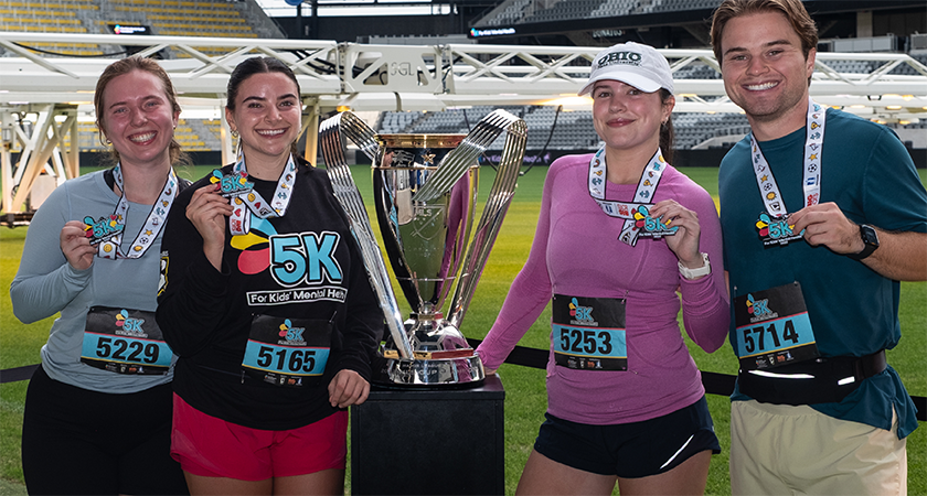 Four participants at a Kids Mental Health Foundation 5K race smiling and holding medals beside a large trophy on a stadium field, wearing race bibs and athletic gear