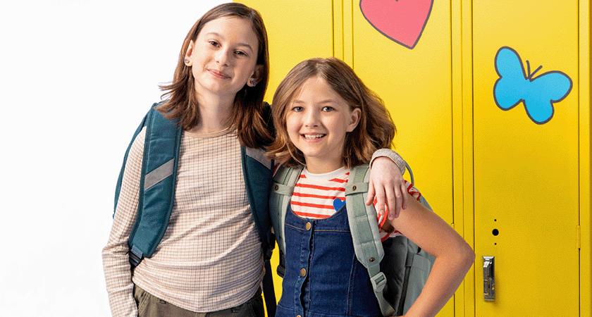 Two smiling school-age girls with backpacks stand in front of bright yellow lockers decorated with a heart and butterfly, symbolizing friendship, confidence, and a positive back-to-school experience.