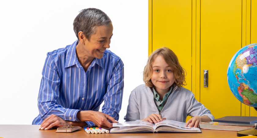 Teacher smiles while helping a young student read a book at a classroom desk, with yellow lockers and a globe in the background, representing early childhood learning and mental health support in education.