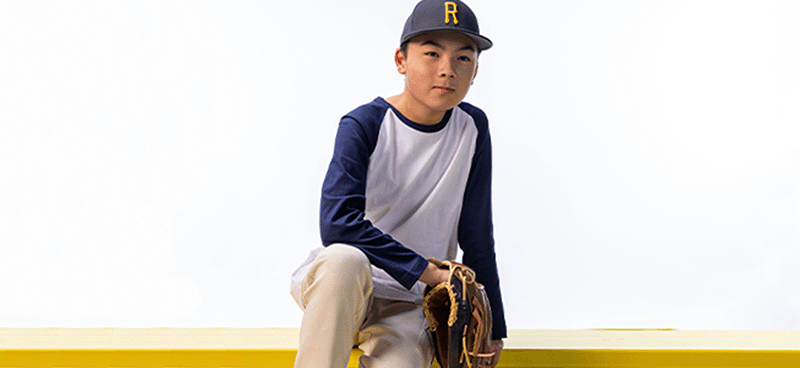 Young baseball player in uniform and cap sits on a yellow bench holding a glove, symbolizing youth resilience, athletic identity, and the importance of mental health support for student athletes.