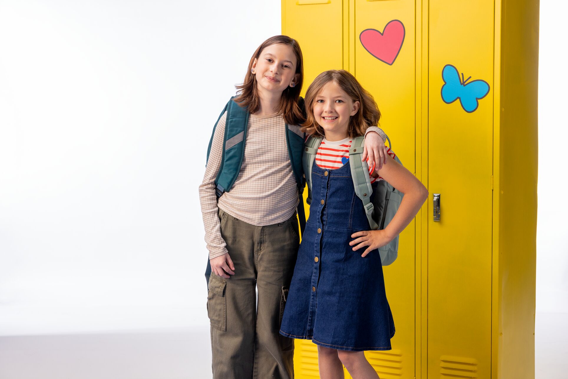 Two smiling elementary school-aged children wearing backpacks stand together in front of bright yellow school lockers decorated with a heart and butterfly, representing a positive back-to-school environment.
