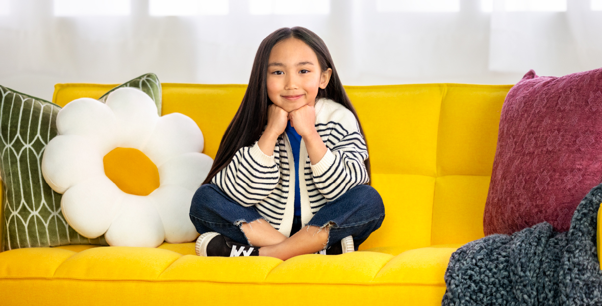 Smiling young girl sits cross-legged on a bright yellow couch surrounded by colorful pillows, representing confidence, comfort, and emotional well-being in a safe space.