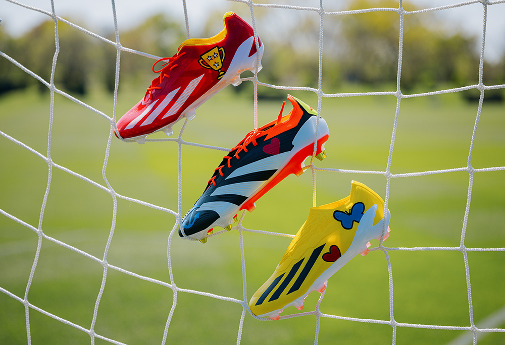 Three colorful soccer cleats featuring heart and butterfly designs hang on a net against a green field background, symbolizing creativity, play, and support for youth mental health.