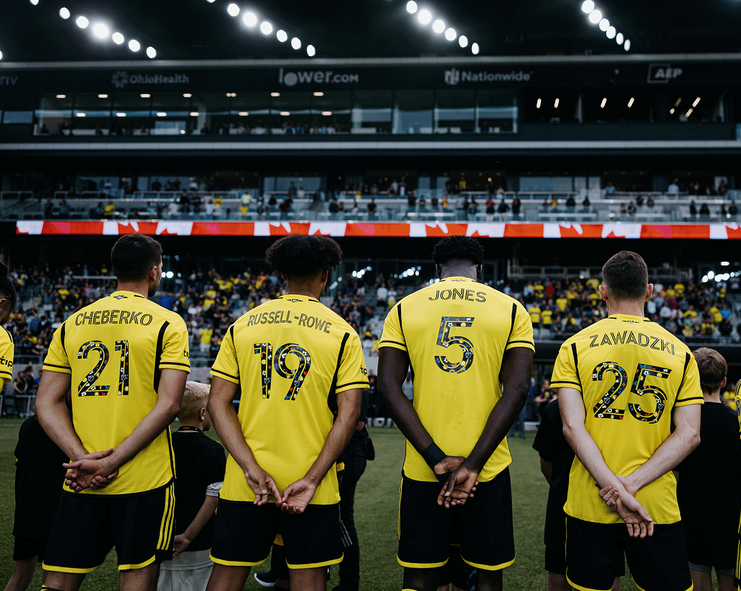 Columbus Crew soccer players Cheberko, Russell-Rowe, Jones, and Zawadzki stand on the field in yellow uniforms before a match at Lower.com Field, facing a packed stadium during pre-game ceremonies.
