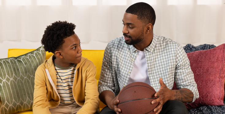 Supportive adult holds a basketball while talking with a young boy on a yellow couch, representing open conversations about bullying prevention, emotional support, and youth mental health.