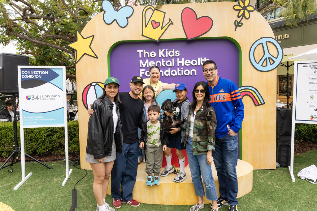 Families and community members smile in front of a colorful Kids Mental Health Foundation photo backdrop featuring peace signs, hearts, and stars at an outdoor event co-hosted with Univision Los Angeles.