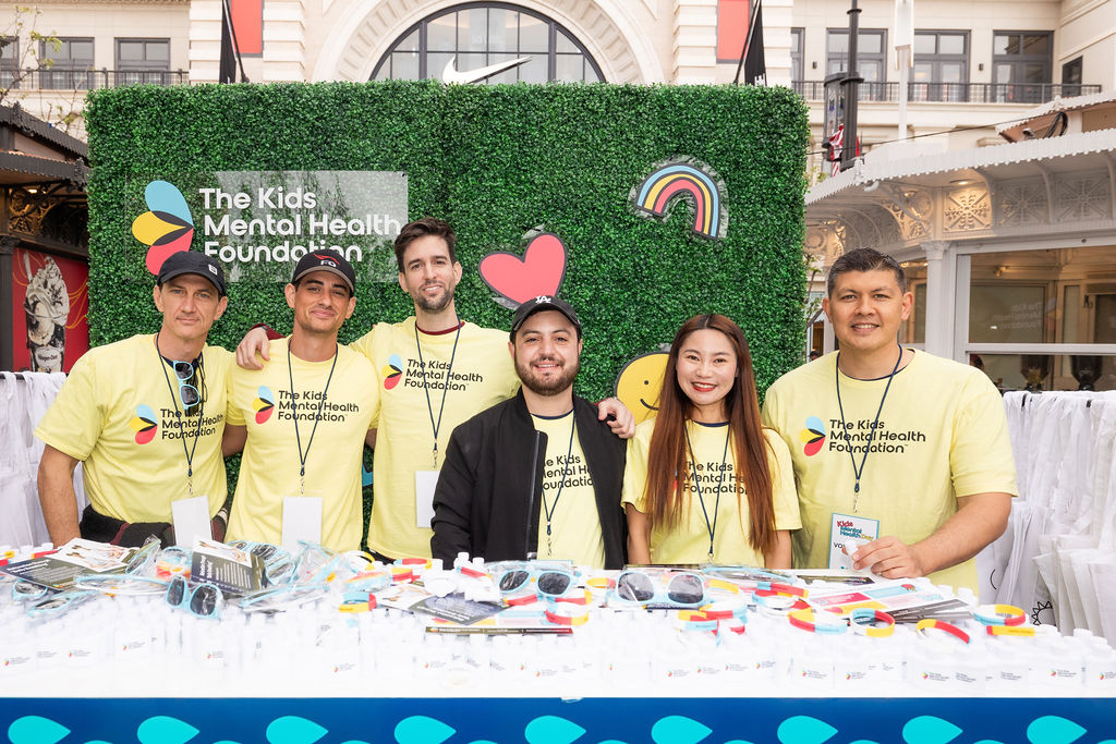 Team of volunteers in yellow Kids Mental Health Foundation shirts stand behind a booth filled with sunglasses, buttons, and informational materials, ready to welcome event attendees.
