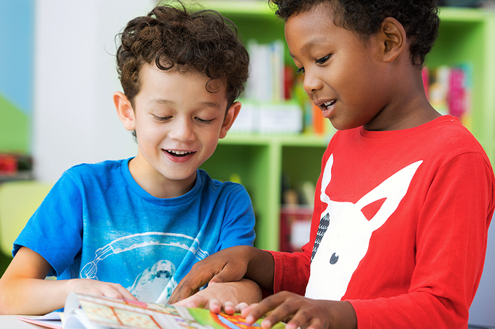 wo young boys smiling and reading a colorful book together in a classroom setting, highlighting peer learning and early childhood literacy in a supportive school environment.