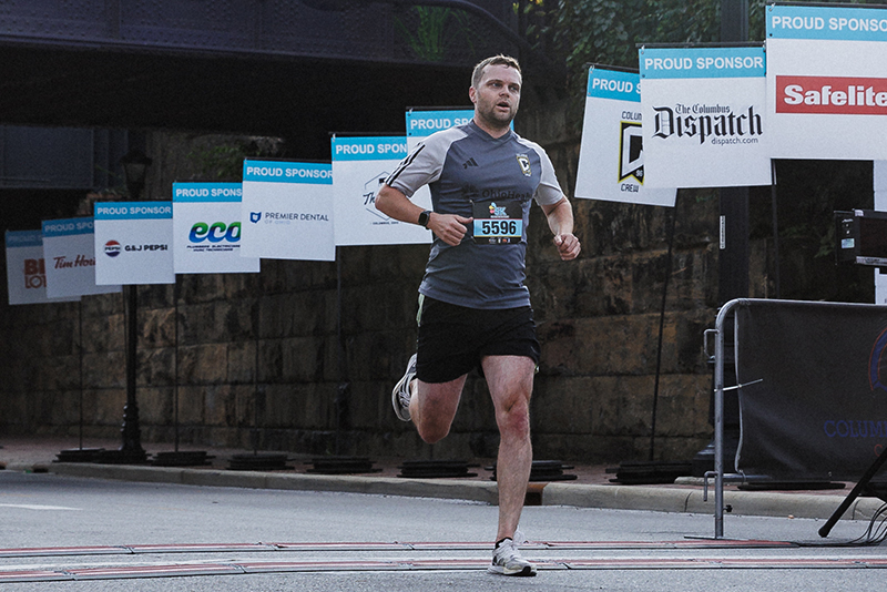 Runner crosses the finish line during the 5K For Kids’ Mental Health race, passing sponsor signage from Columbus Crew partners, supporting youth mental health awareness.