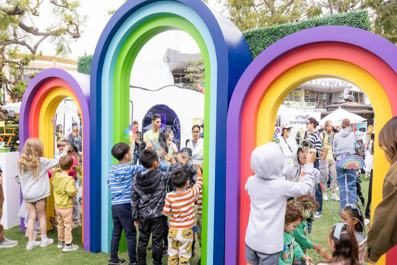 Children interact with rainbow-colored arch installations and draw on glass panels at an outdoor mental health event designed to spark creativity and connection.