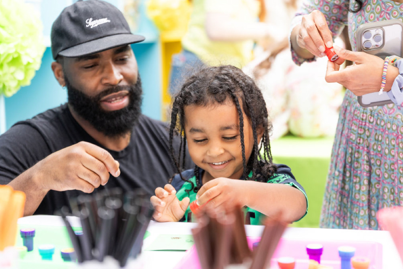A young child smiles while working on a hands-on art project with an adult during a creative mental health event focused on bonding and emotional expression.