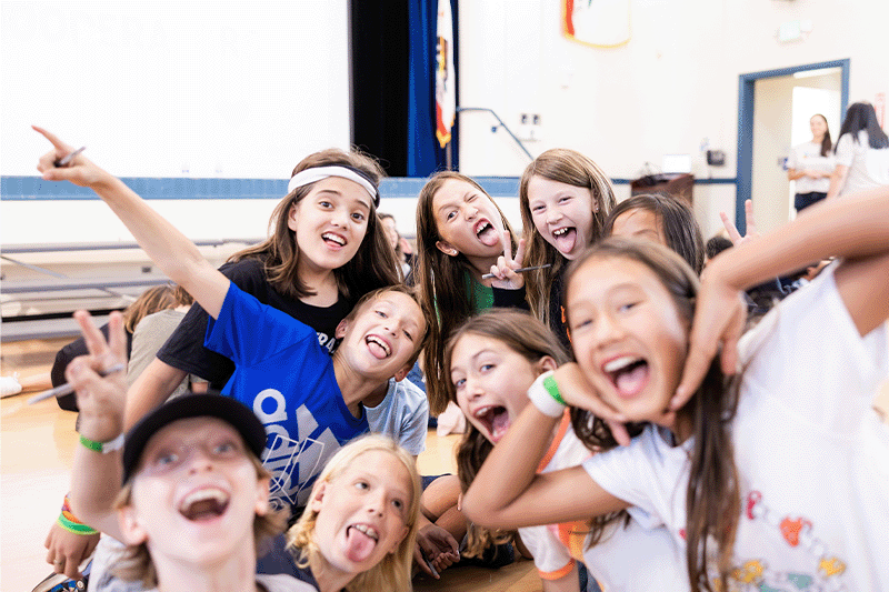 A diverse group of preteens laugh and pose together inside a school gym, celebrating friendship and community during a mental health awareness event.