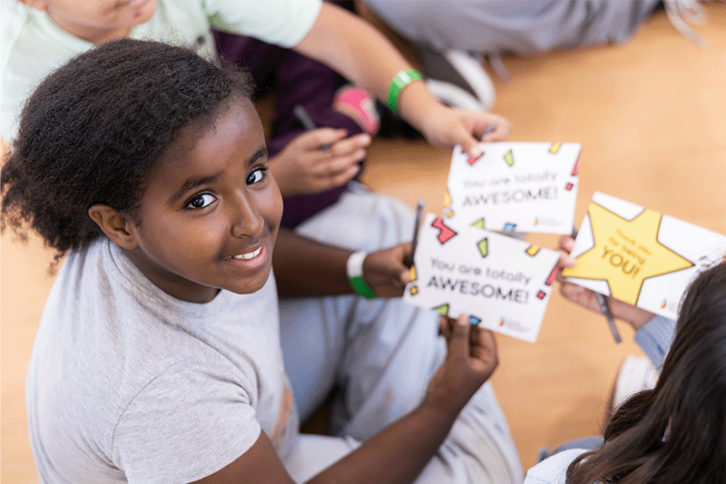 A student smiles while holding a card that says "You are totally AWESOME!" during a school activity focused on building confidence and peer encouragement.