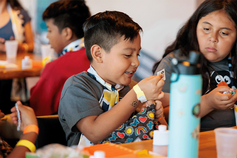 Smiling young boy explores a colorful sensory bag during a hands-on mental wellness activity, surrounded by other children at a community event focused on emotional health and self-expression.