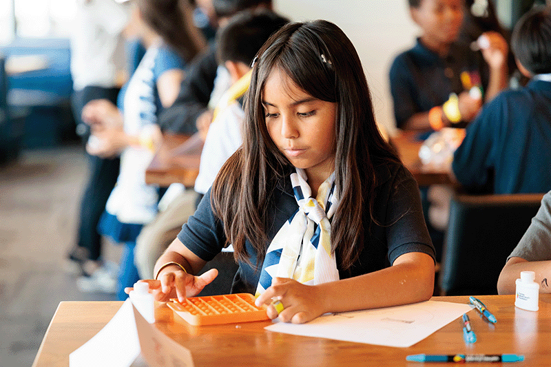 Focused young girl participates in a hands-on learning activity using a sensory fidget tool during a mental health workshop, surrounded by classmates engaged in creative expression and emotional regulation exercises.