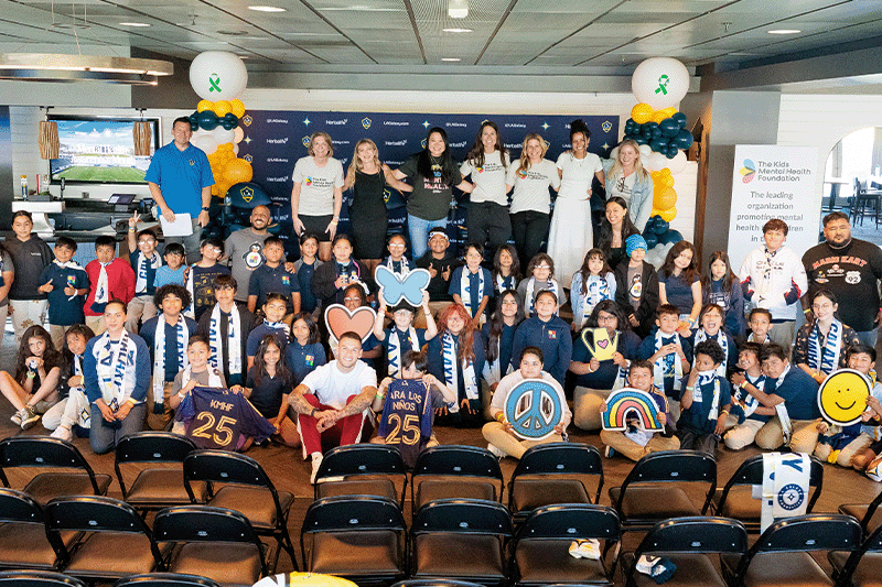 LA Galaxy soccer player Christian Ramirez poses with students, educators, and Kids Mental Health Foundation staff at a school mental wellness event. The group holds playful icons like hearts and rainbows, celebrating youth mental health awareness and community support.