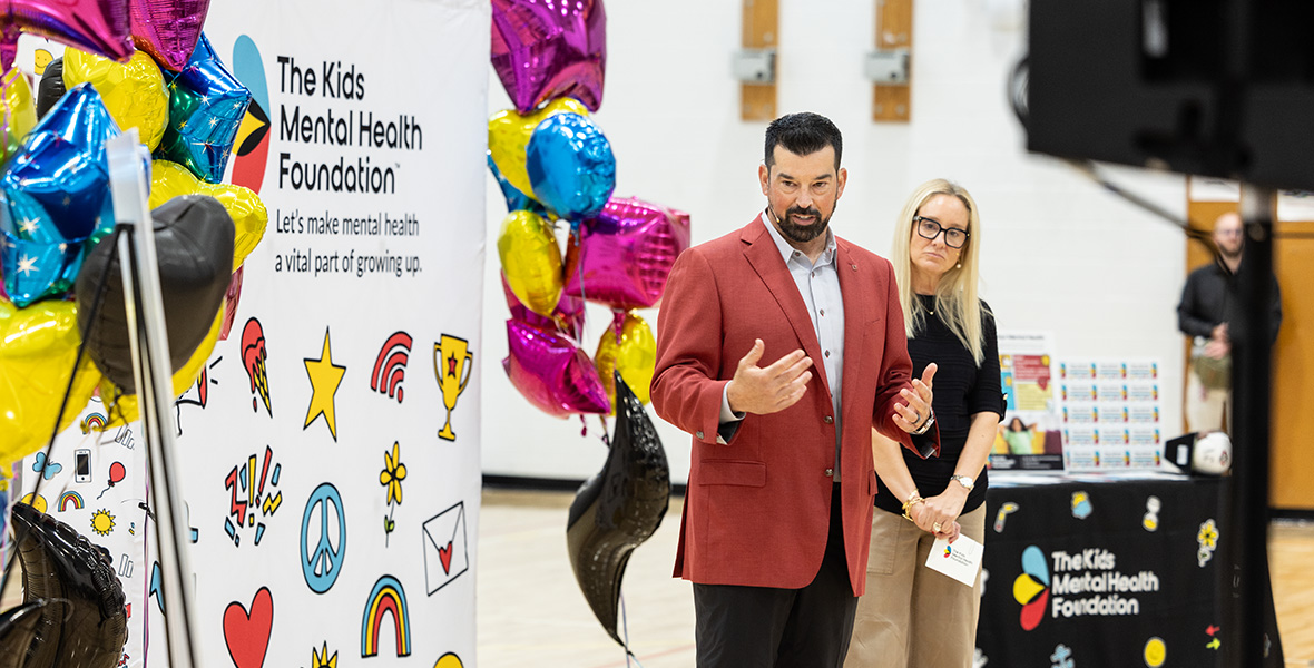 Ryan Day interviene en un acto de la Fundación para la Salud Mental Infantil, junto a un expositor de la fundación y unos globos de colores, en el gimnasio de un colegio.
