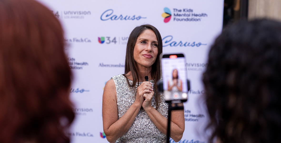 La actriz y activista Soleil Moon Frye interviene en un acto de la Kids Mental Health Foundation, de pie frente a un telón de fondo con el logotipo de la organización, mientras los asistentes la graban con sus móviles, con el fin de sensibilizar sobre el bienestar mental de los jóvenes.