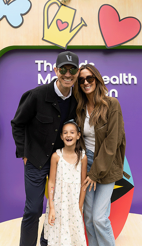 Angel Carter Conrad posa junto a su esposo y su hija frente a un colorido telón de fondo de la Fundación para la Salud Mental Infantil, sonriendo juntos en un evento comunitario sobre salud mental que celebra los lazos familiares y el bienestar emocional.