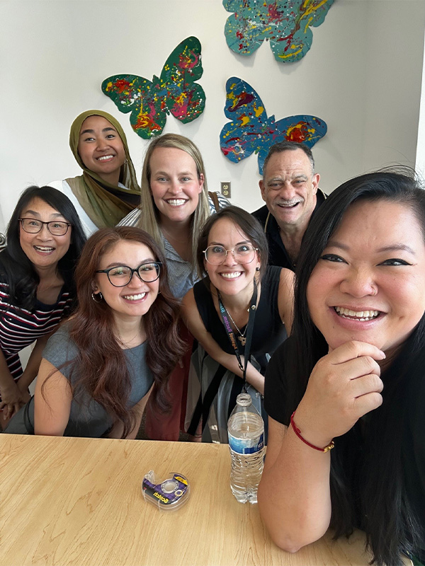 La actriz Anna Cathcart sonríe junto a un grupo de miembros del equipo de The Kids Mental Health Foundation, posando frente a una colorida obra de arte con mariposas y celebrando la defensa de la salud mental de los jóvenes.