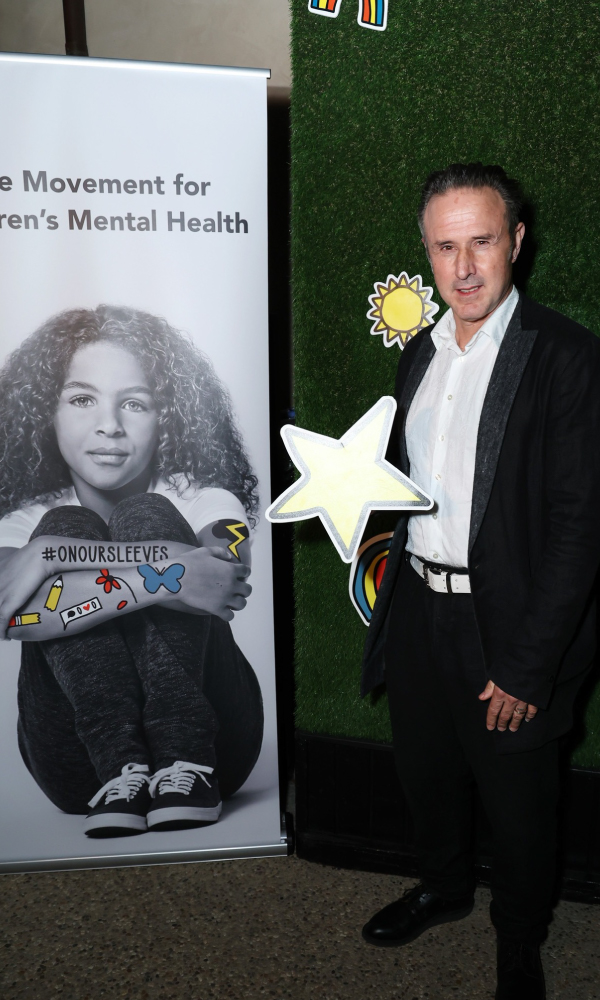 David Arquette posa junto a un expositor que promueve la salud mental infantil, sosteniendo una estrella recortada que brilla, en un acto de la Fundación para la Salud Mental Infantil centrado en la sensibilización y el apoyo.
