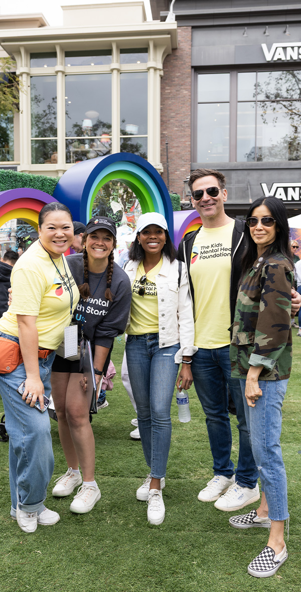 La periodista y activista Lisa Ling posa junto a un grupo de voluntarios en un acto de la Fundación para la Salud Mental Infantil, sonriendo frente a unos arcos y escaparates de colores vivos con motivos del arcoíris.