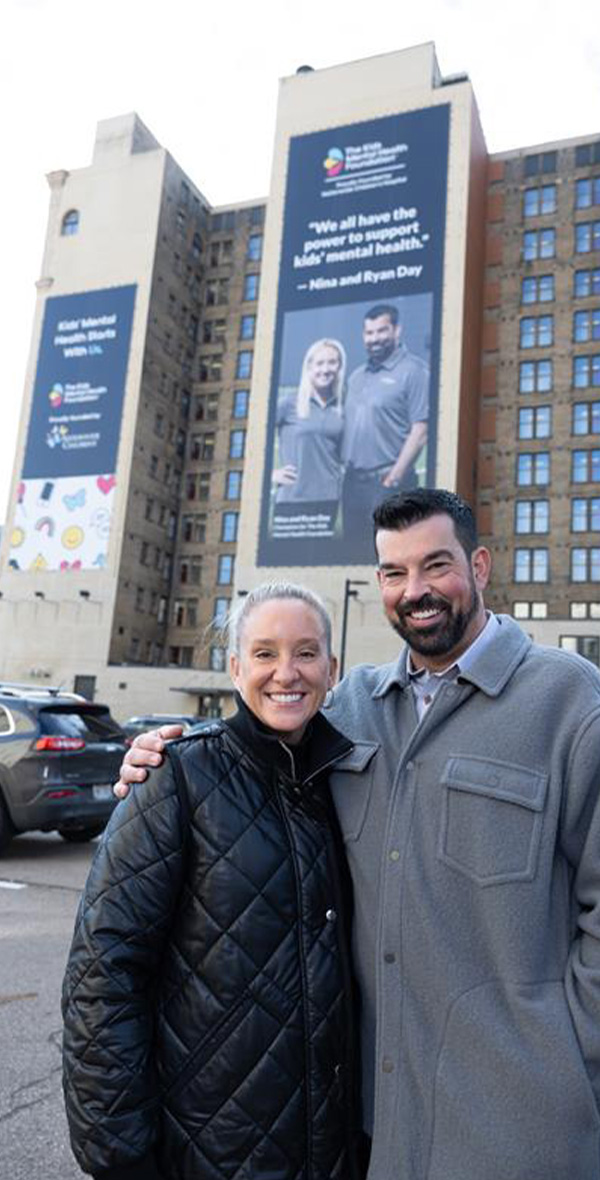 Nina y Ryan Day posan sonrientes frente a un edificio del centro de la ciudad en el que se exhibe una gran pancarta de la Fundación para la Salud Mental Infantil con su foto y la siguiente cita: «Todos tenemos el poder de apoyar la salud mental de los niños».