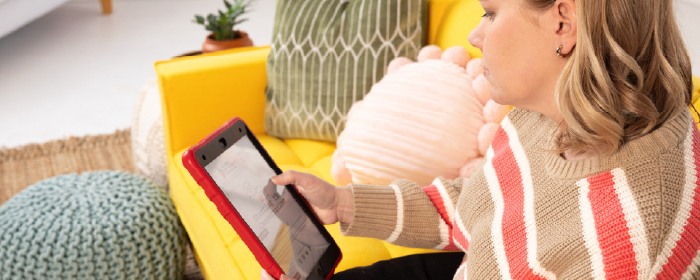 Un padre está sentado en un sofá amarillo leyendo en una tableta, rodeado de mullidos cojines, promoviendo el acceso a recursos digitales de salud mental y el apoyo a las familias en el hogar.