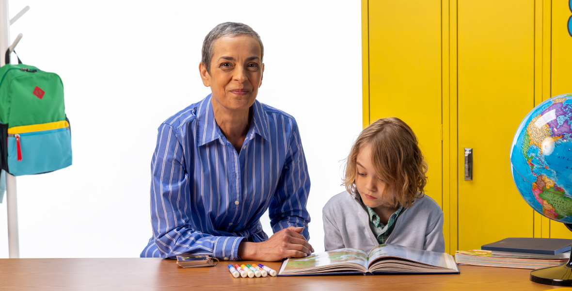 Teacher sits beside a young student reading a book at a classroom desk, with bright yellow lockers, a globe, and school supplies in the background, highlighting early childhood learning and mental health support in education.