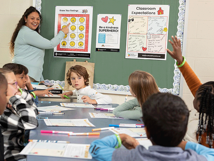 Una profesora de primaria dirige una actividad en el aula sobre la conciencia emocional utilizando una tabla titulada «Conoce tus sentimientos», mientras los alumnos trabajan con las fichas y levantan la mano, fomentando así el aprendizaje socioemocional a través del kit para profesores de la Fundación para la Salud Mental Infantil.
