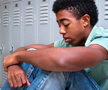 Un adolescente sentado en el suelo con los ojos cerrados y los brazos alrededor de las rodillas frente a las taquillas del colegio, en representación de los problemas de salud mental entre los jóvenes que se ponen de manifiesto en las encuestas de los CDC.