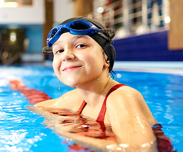 Una niña sonriente, vestida con bañador y gorro de baño, descansa junto al borde de una piscina, promoviendo la actividad física como un microhábito que favorece el bienestar mental de los niños.
