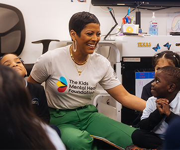 Tamron Hall, vestida con una camiseta de la Fundación para la Salud Mental Infantil, sonríe e interactúa con un grupo de jóvenes estudiantes durante una visita a las aulas, con el fin de fomentar la concienciación sobre la salud mental y el sentido de comunidad.